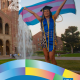 Ajana Orozco ’21 stands in front of UCLA’s Royce Hall and fountain wearing graduation regalia, holding a transgender pride flag overhead. Text identifies her field of study as Gender Studies and includes CCCP’s 25 Years and UCLA logos.