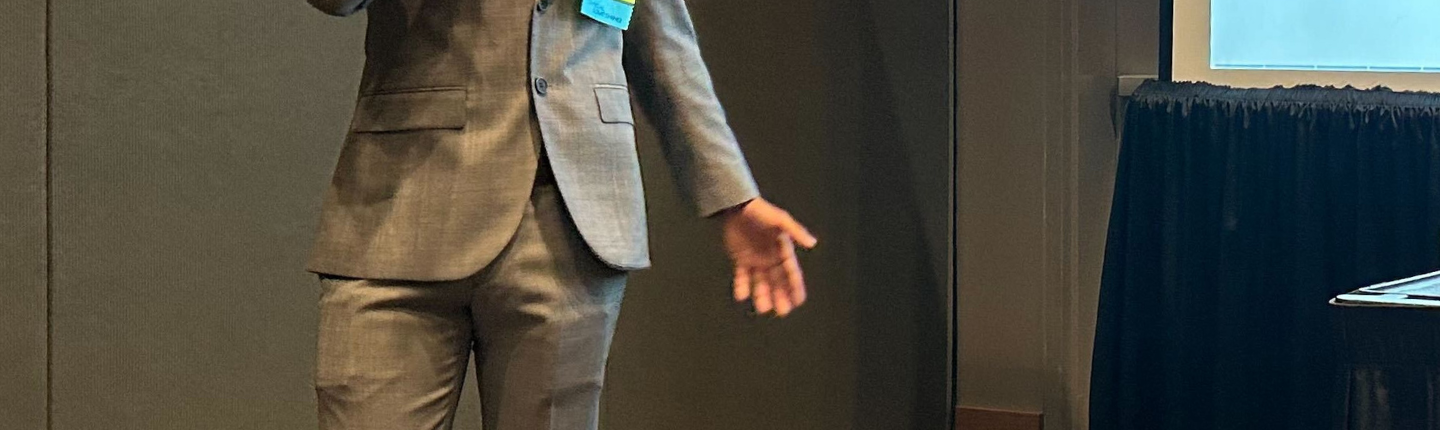Nate Hoffman, UCLA Communication class of 2024, speaks into a microphone beside a presentation screen in a CCCP 25th anniversary–branded alumni graphic.