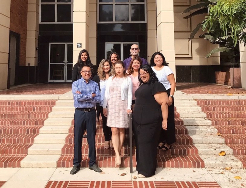 Eight staff members pose together on brick steps outside a UCLA campus building. Three people stand in the front row while the remaining group stands behind them near the building entrance.