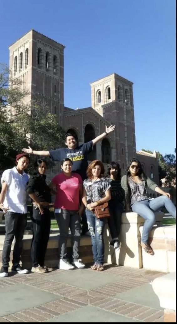 Seven students pose outdoors in front of UCLA’s Royce Hall on a sunny day. One student stands behind the group with both arms raised, while another sits on a low wall at the right. The building’s twin towers and a clear blue sky appear in the background.