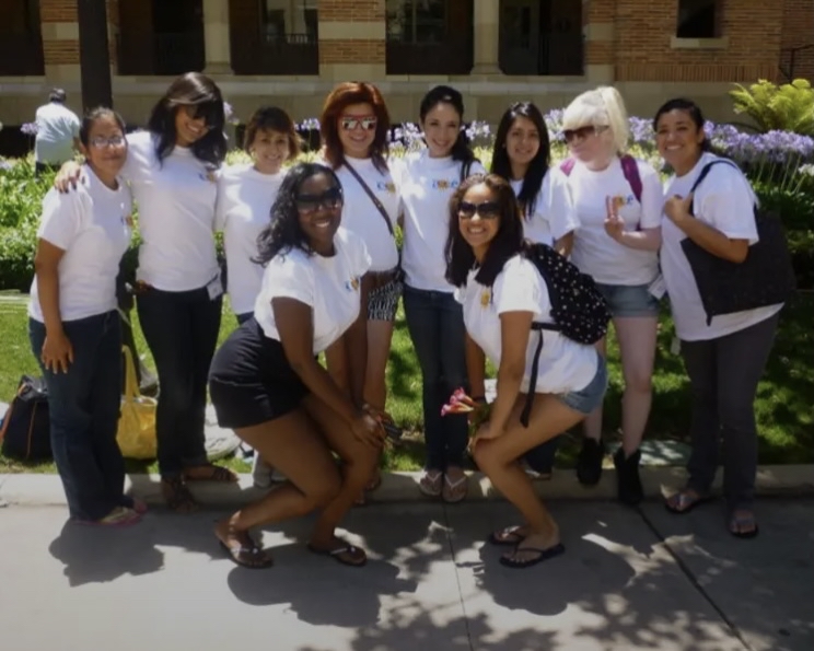 Nine students wearing white T-shirts pose together outdoors on a UCLA campus walkway. Two students crouch in front while the others stand behind them, surrounded by grass, flowers, and a brick campus building.