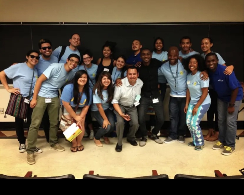 A diverse group of students and staff wearing mostly light blue shirts pose together at the front of a classroom. Several participants wear name badges, and a blackboard fills the background.