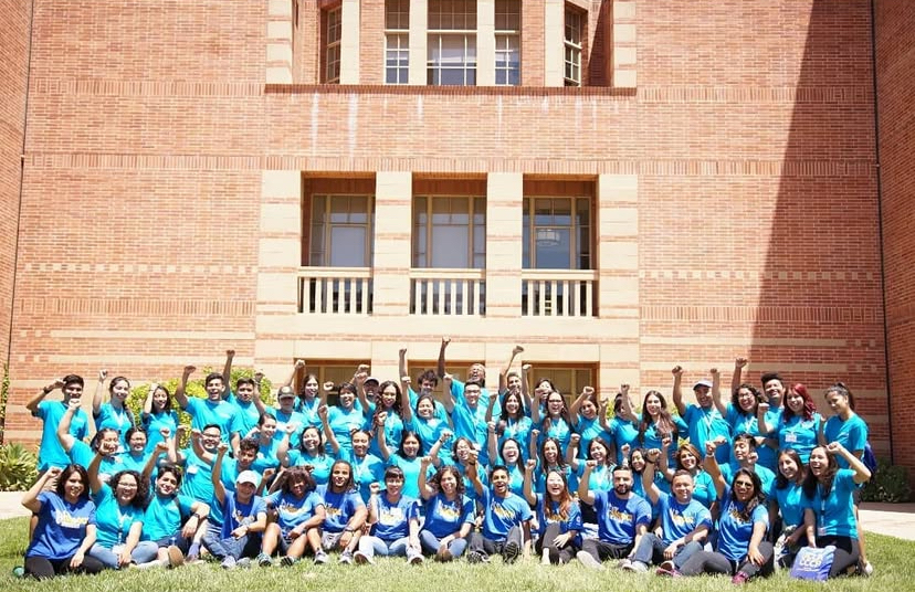 A large group of students and staff wearing blue and turquoise shirts pose on a lawn in front of a UCLA brick building. Many participants raise their fists or arms in celebration.