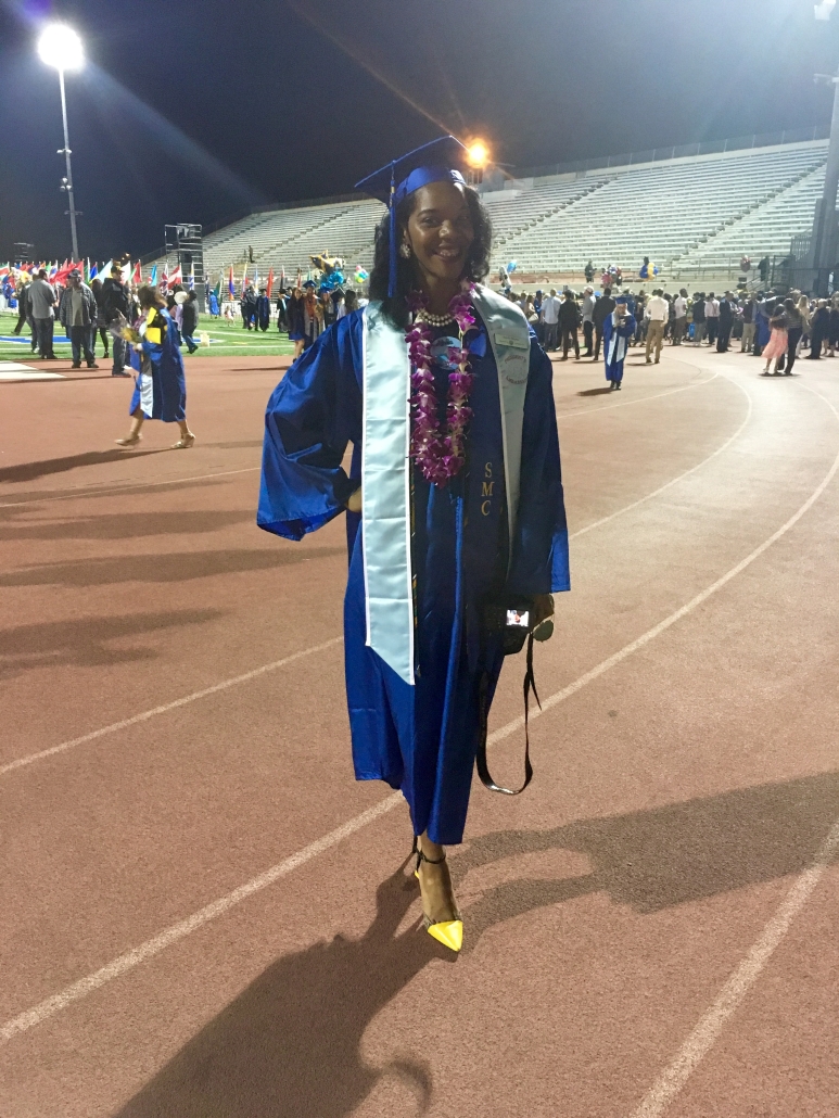 Aurelia poses on a stadium track after graduating from Santa Monica College, wearing a blue cap and gown, honor stoles, and a purple flower lei.