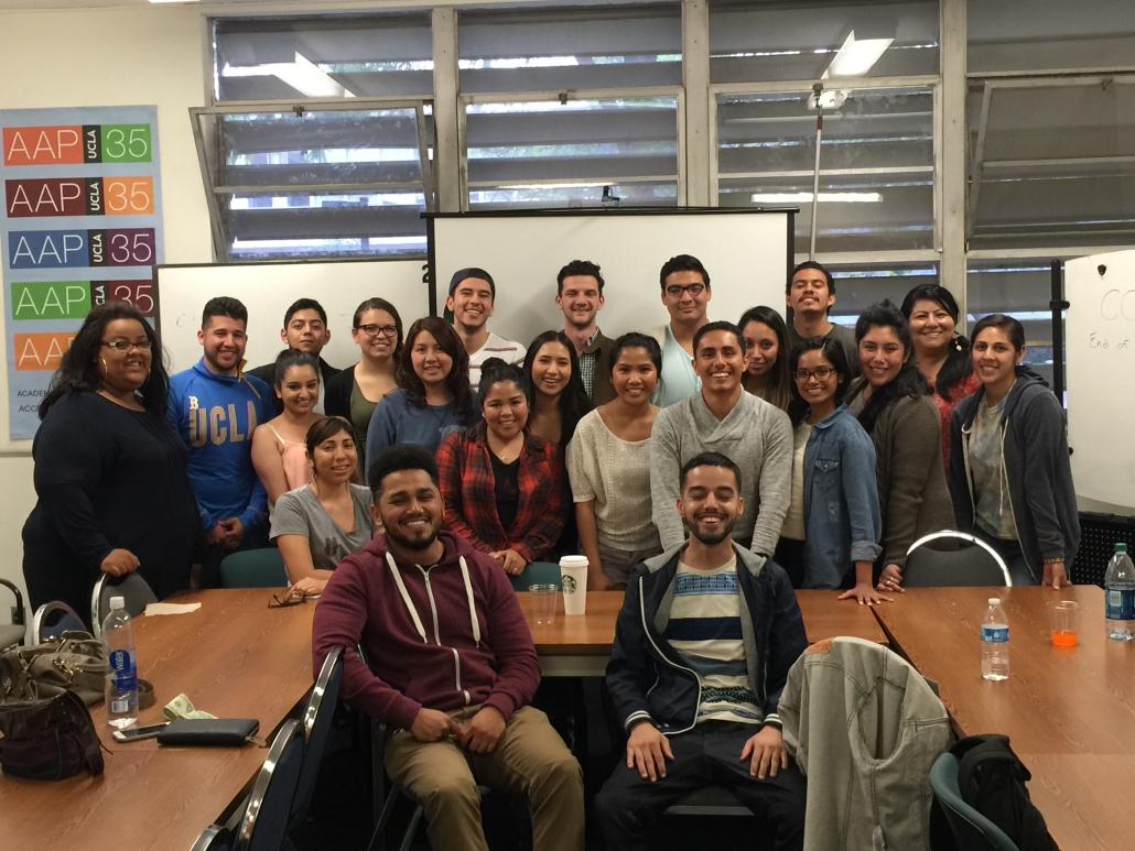 A large group of students and staff pose together inside a classroom around conference tables, with whiteboards, windows, and AAP UCLA 35 posters in the background.