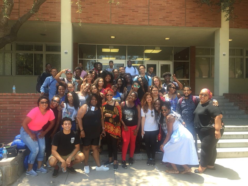 A large group poses outdoors on the steps of a campus building on a sunny day, with several participants standing in rows and one person crouching in front.