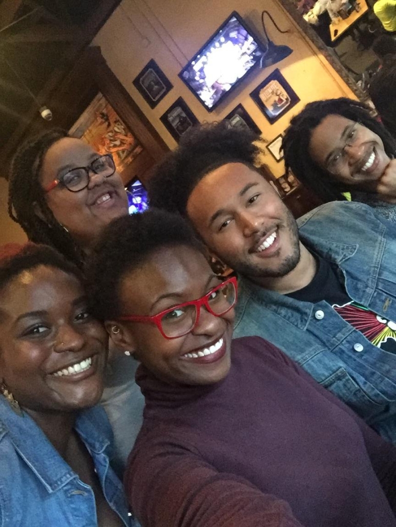Five people smile for a close-up group selfie inside a restaurant, with framed wall art and television screens visible behind them.