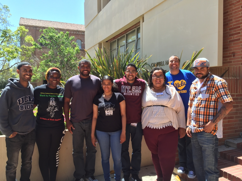 Eight people pose outdoors in front of Campbell Hall at UCLA, with campus buildings, trees, and tall green plants in the background.