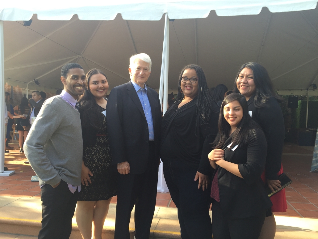 Six people pose together beneath a large outdoor event tent, including Gene Block in a dark suit at the center and guests dressed in business attire.
