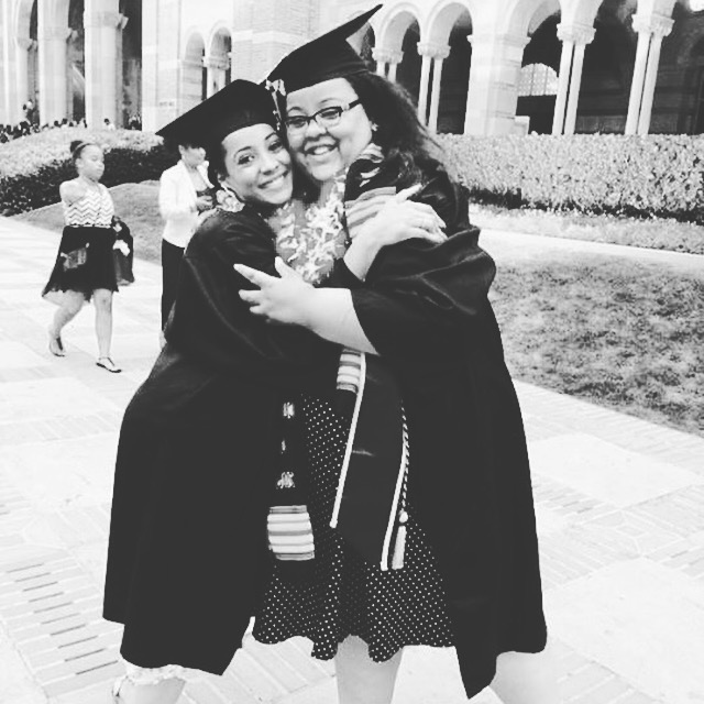 Black-and-white photo of two graduates in caps and gowns embracing and smiling outdoors near a campus building with arched walkways.