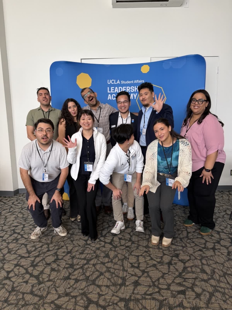 Ten participants pose playfully in front of a blue UCLA Student Affairs Leadership Academy backdrop, with several people waving or making expressive faces.