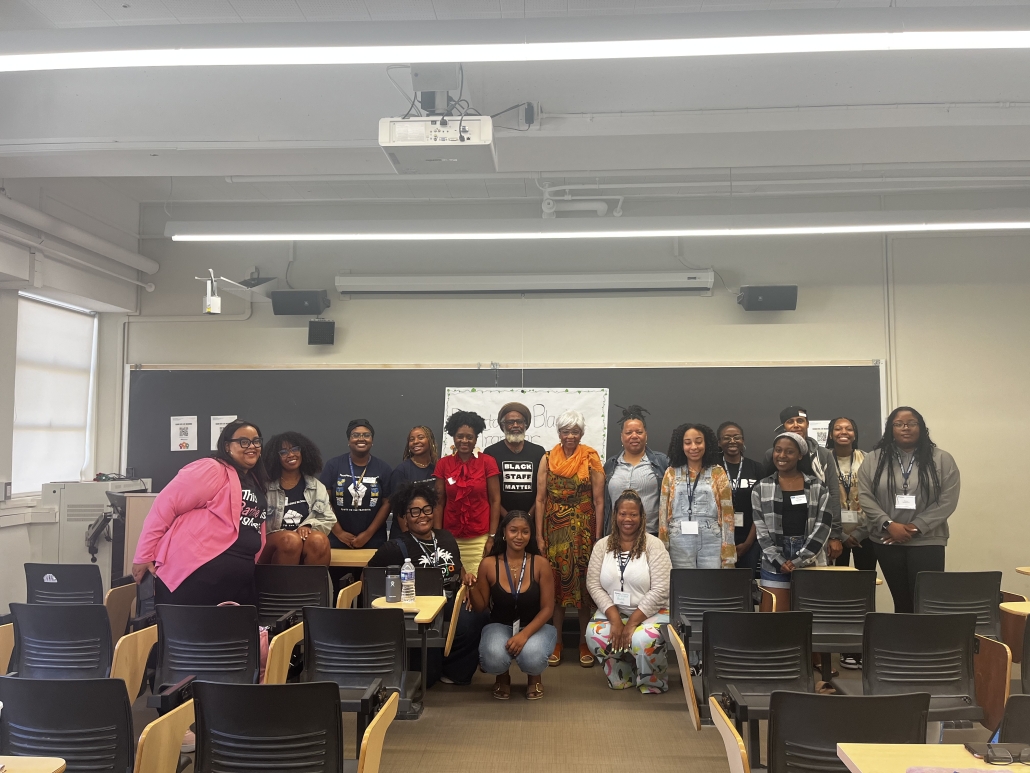 A group of participants pose together at the front of a classroom beneath a sign referencing Black staff, with rows of lecture chairs and a large chalkboard visible.