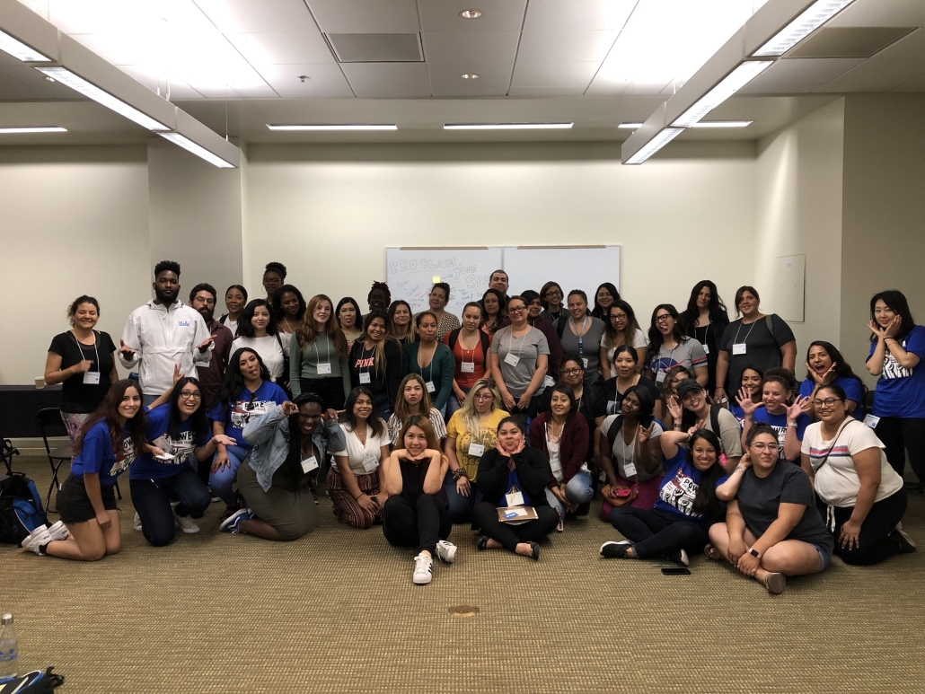 A large group of students and staff posing playfully inside a classroom, with several participants wearing blue “Power to the Transfer” shirts and name badges.