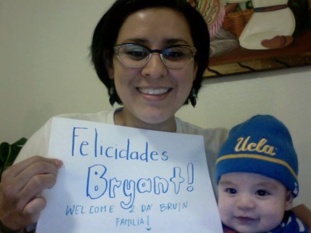 Christine Vega smiles beside a baby wearing a UCLA beanie while holding a handwritten sign that reads, “Felicidades Bryant! Welcome 2 da Bruin familia!”