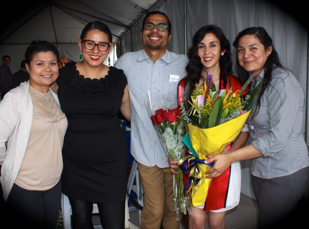 Christine Vega, wearing a UCLA graduation stole and holding bouquets of flowers, poses with four supporters at a graduation celebration.