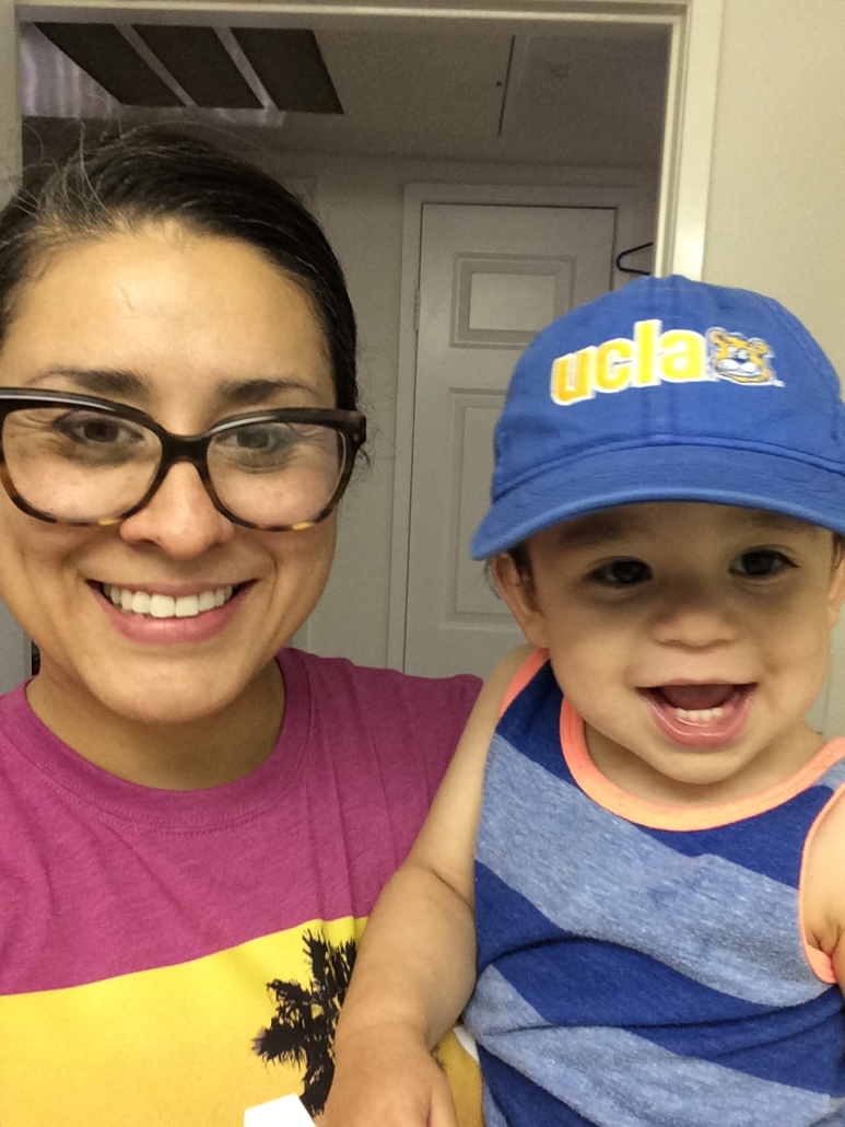 Christine Vega smiles while holding a laughing baby wearing a blue UCLA cap.