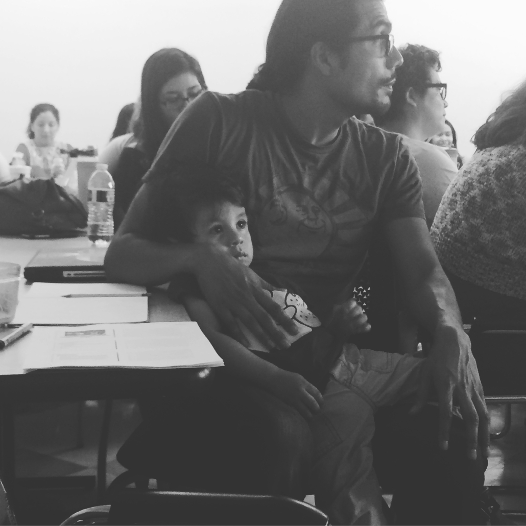 Black-and-white photo of a man seated at a classroom table with a young child resting against him during a group gathering.