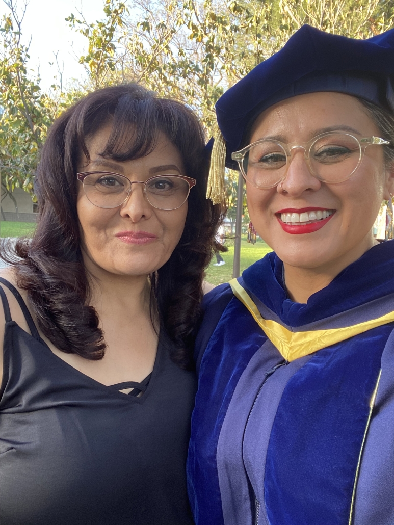 Christine Vega, wearing UCLA doctoral regalia, takes a graduation-day selfie with a woman outdoors.