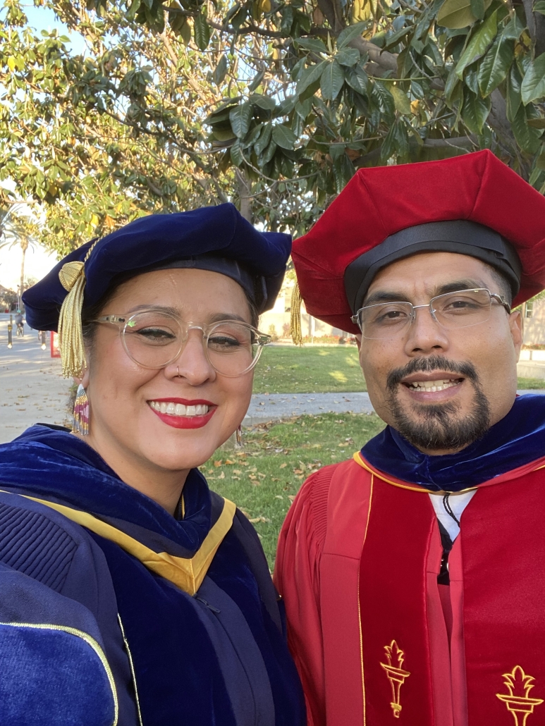 Christine Vega and a colleague smile for a selfie while wearing doctoral regalia at an outdoor graduation ceremony.