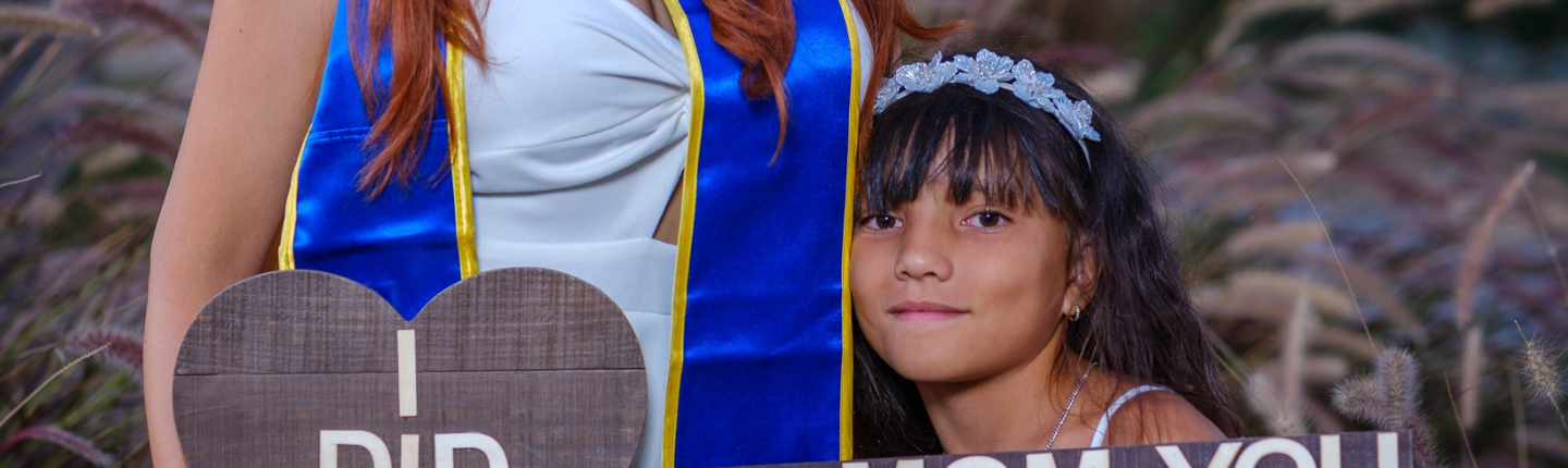 Brenda Coronel, UCLA Sociology class of 2022 and M.A. in Social Welfare class of 2026, poses in graduation regalia beside a child holding a celebratory sign.