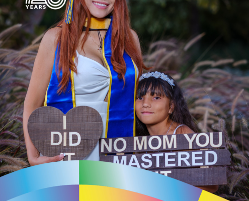 Brenda Coronel, UCLA Sociology class of 2022 and M.A. in Social Welfare class of 2026, poses in graduation regalia beside a child holding a celebratory sign.