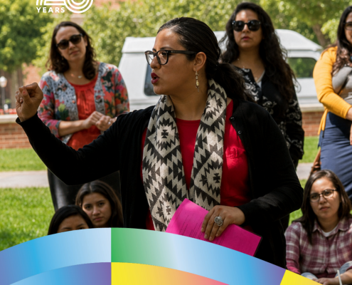 CCCP 25th anniversary profile graphic featuring Christine Vega speaking outdoors to a group of students while holding a bright pink folder. She wears glasses, a red top, black cardigan, and a black-and-white patterned scarf. The white CCCP 25 Years logo appears at the top left, colorful curved ribbon graphics frame the bottom, and the UCLA logo appears at the lower right. Text reads: “Christine Vega ’07, Ph.D. ’19, Women & Gender Studies and Chicana/o Studies, Education and Comparative Race and Ethnic Studies.”