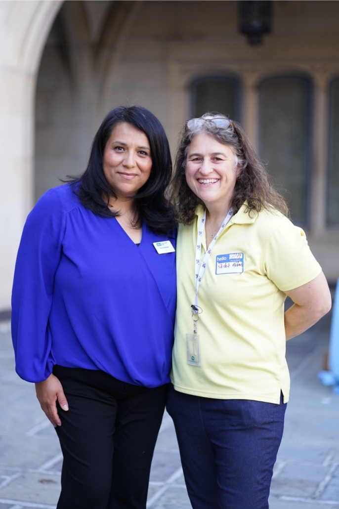 Carina Salazar poses with another woman in a UCLA courtyard. Carina wears a blue blouse and name badge, while the other woman wears a pale yellow polo shirt and event lanyard.
