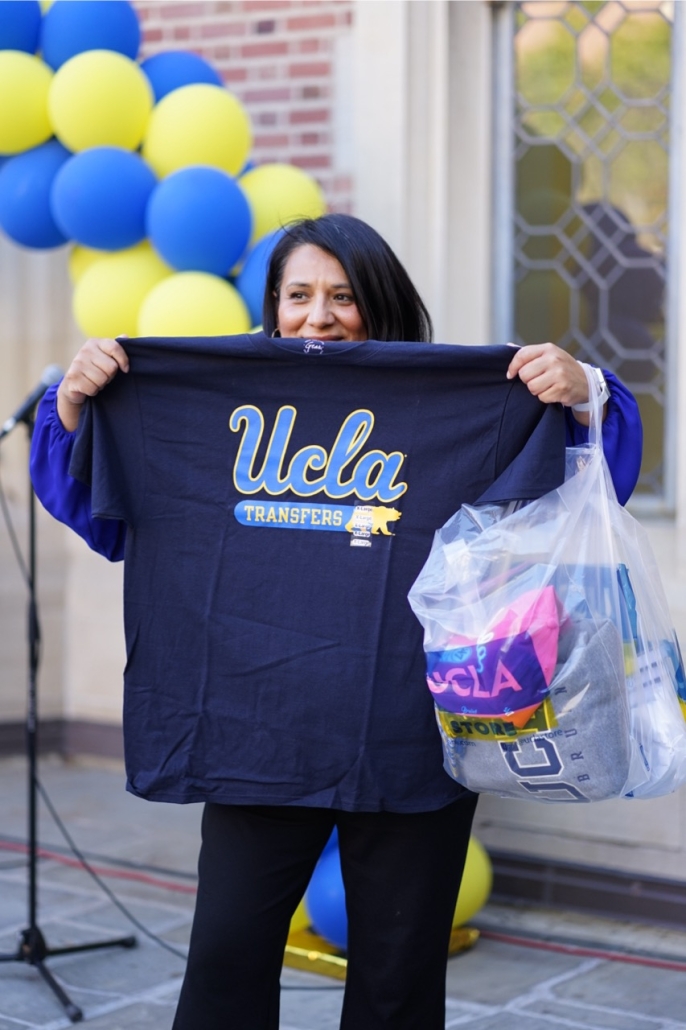 Carina Salazar holds up a navy UCLA Transfers T-shirt while carrying a clear bag filled with UCLA merchandise at an outdoor campus event decorated with blue and yellow balloons.