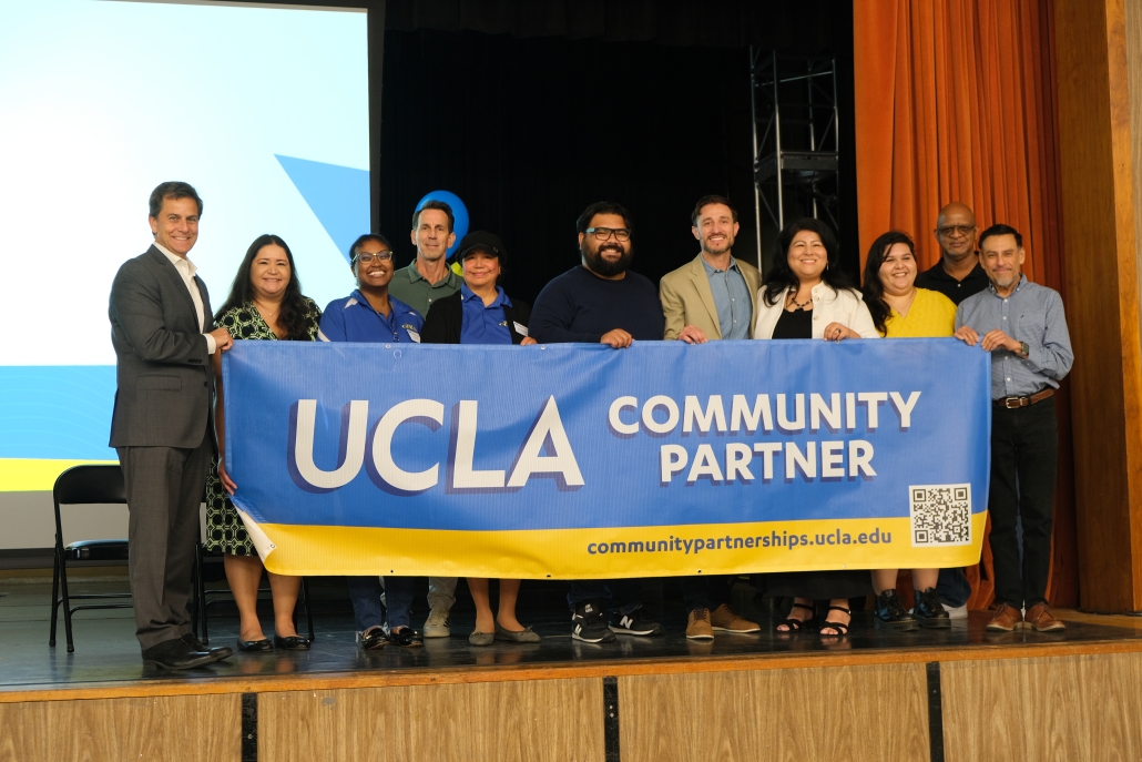 Jessica Hall, third from the left, joins UCLA staff and community partners onstage as they hold a large blue-and-gold “UCLA Community Partner” banner.