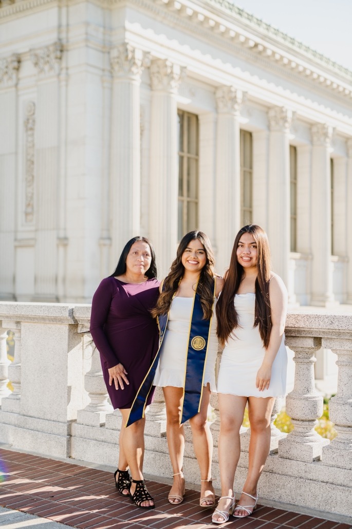 Ariana Reyes poses in a white dress and blue-and-gold UCLA graduation stole with two women on a campus terrace in front of a columned building.