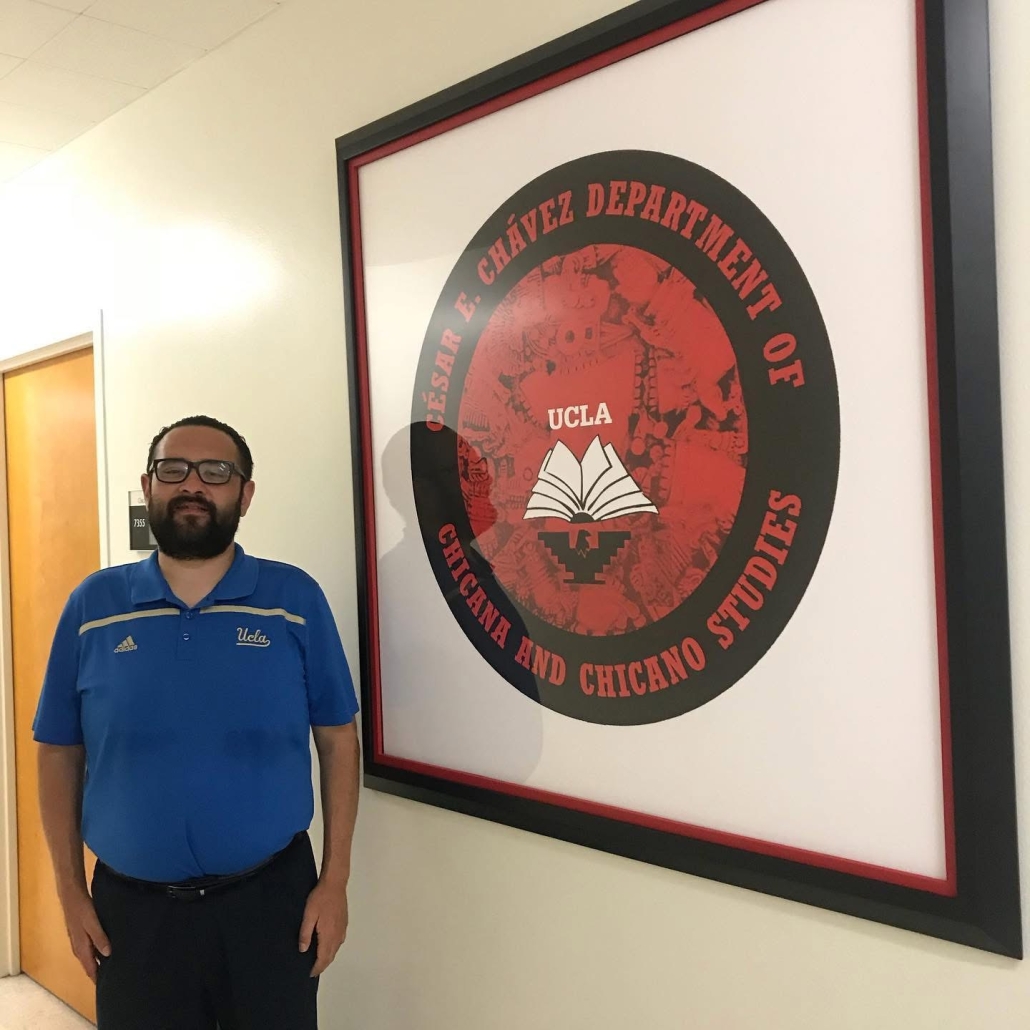 José Del Real Viramontes stands in a hallway beside a large framed sign for UCLA’s César E. Chávez Department of Chicana and Chicano Studies. He wears glasses and a blue UCLA polo shirt.