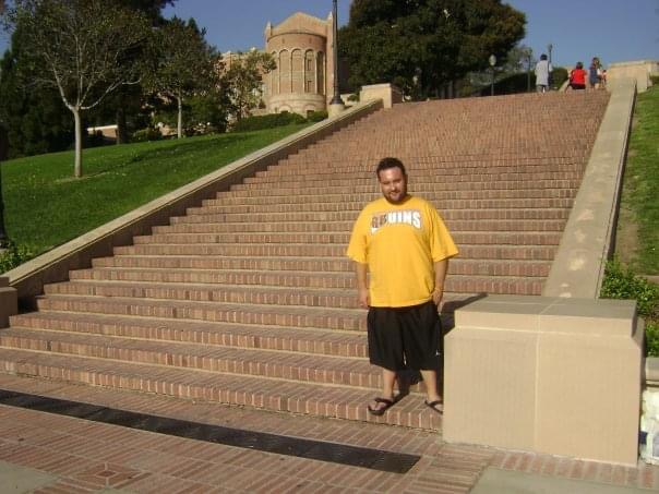 José Del Real Viramontes stands near the bottom of a broad brick staircase on the UCLA campus, wearing a yellow Bruins T-shirt, black shorts, and sandals. Campus buildings, trees, and several people appear at the top of the steps.