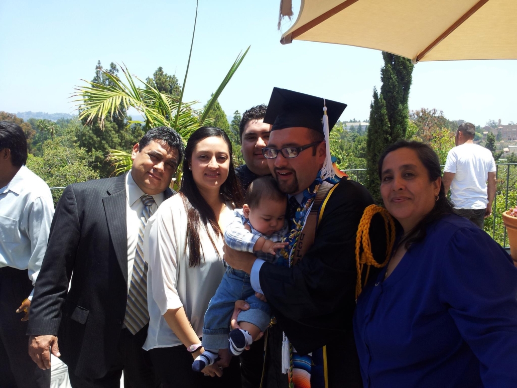 José Del Real Viramontes, wearing a graduation cap and gown, holds a baby while posing outdoors with several relatives and friends. Trees, campus buildings, and other guests appear in the background.