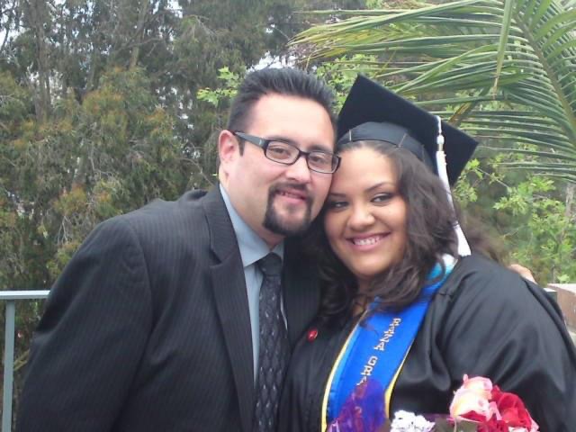 José Del Real Viramontes poses closely with a graduate in a cap and gown who is holding a bouquet of flowers. Both smile at the camera with trees and greenery behind them.