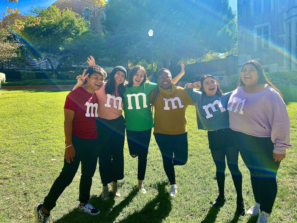 Jessica Hall, fourth from the left, poses with five students wearing colorful shirts decorated with large lowercase “m” letters on a sunny UCLA lawn.