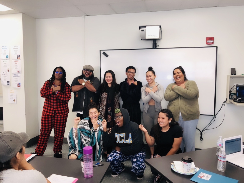 A group of nine students pose together in a classroom in front of a large whiteboard. Several cross their arms over their chests while others laugh and make playful gestures. Desks, notebooks, laptops, water bottles, and student profile posters are visible around the room.