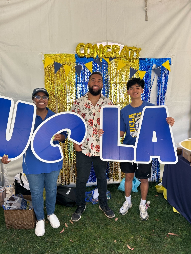 Jessica Hall, wearing glasses and standing on the left, smiles with two others as they hold oversized UCLA letters in front of a blue-and-gold graduation backdrop.
