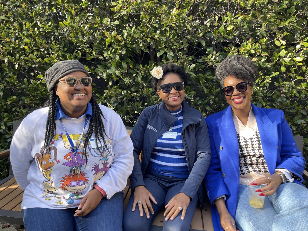 Jessica Hall, seated on the left, smiles with two women on an outdoor bench in front of green foliage.