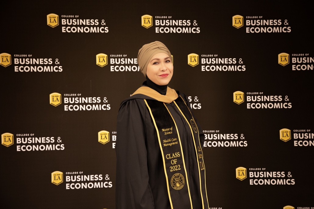 Villy B. poses in a black graduation gown, gold-trimmed stole, and taupe headwrap in front of a Cal State LA College of Business and Economics backdrop. Her stole reads “Master of Science, Health Care Management, Class of 2022.”
