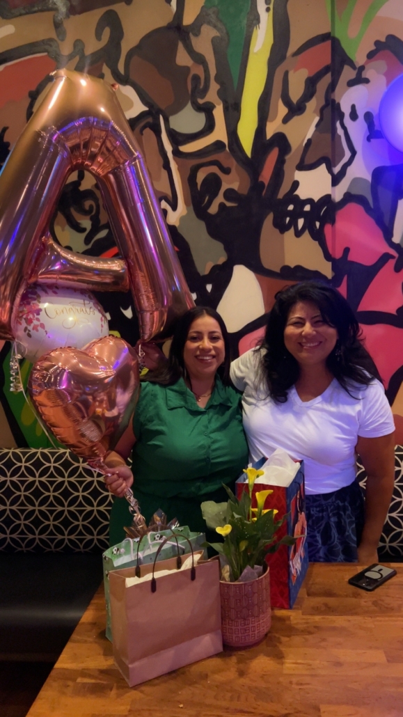 Aranzazu and Claudia Salcedo smile beside congratulatory balloons, flowers, and gift bags during a celebration.