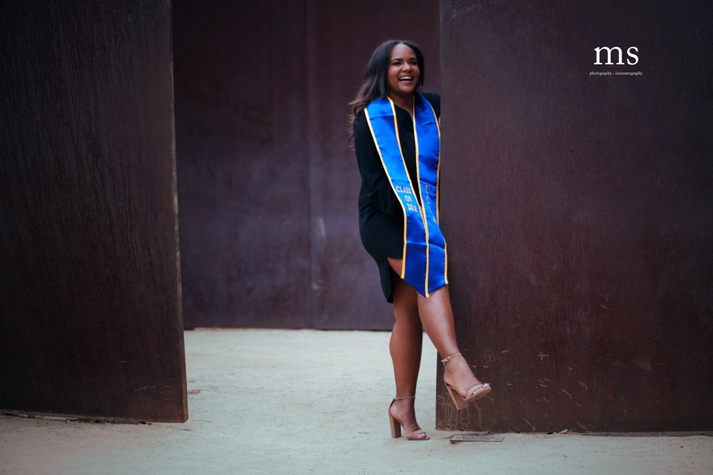 Cynamon Mantley ’18 laughs while posing between large rust-colored metal panels, wearing a black dress and a blue-and-gold UCLA graduation stole.