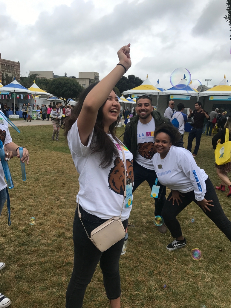 Cynamon Mantley laughs and raises one arm while participating in an outdoor UCLA event with colleagues, tents, and floating bubbles in the background.