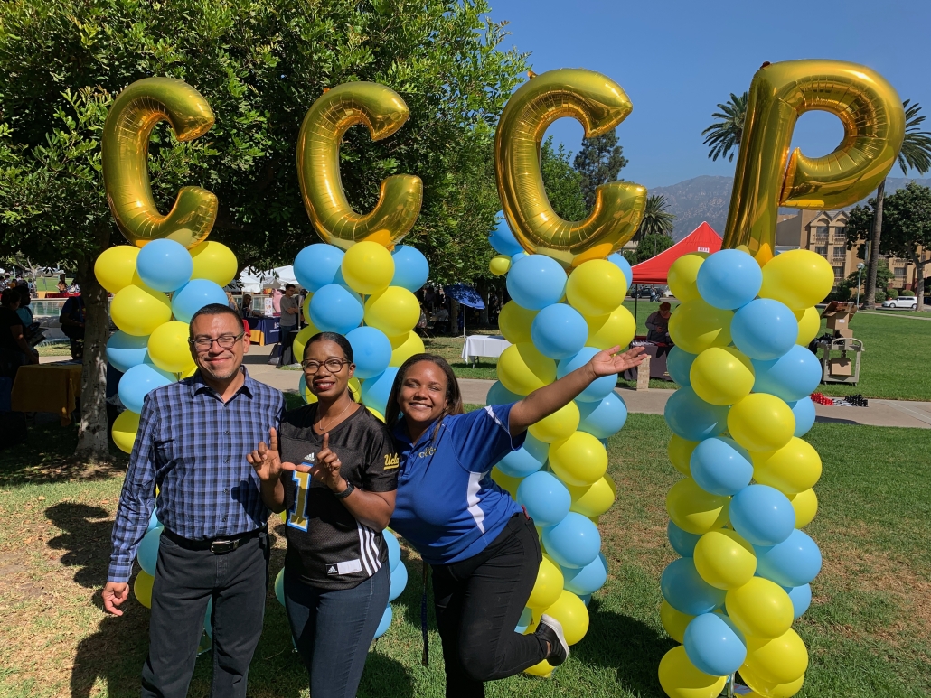 Cynamon Mantley poses with two colleagues beneath large gold CCCP balloon letters supported by blue-and-yellow balloon columns at an outdoor UCLA event.
