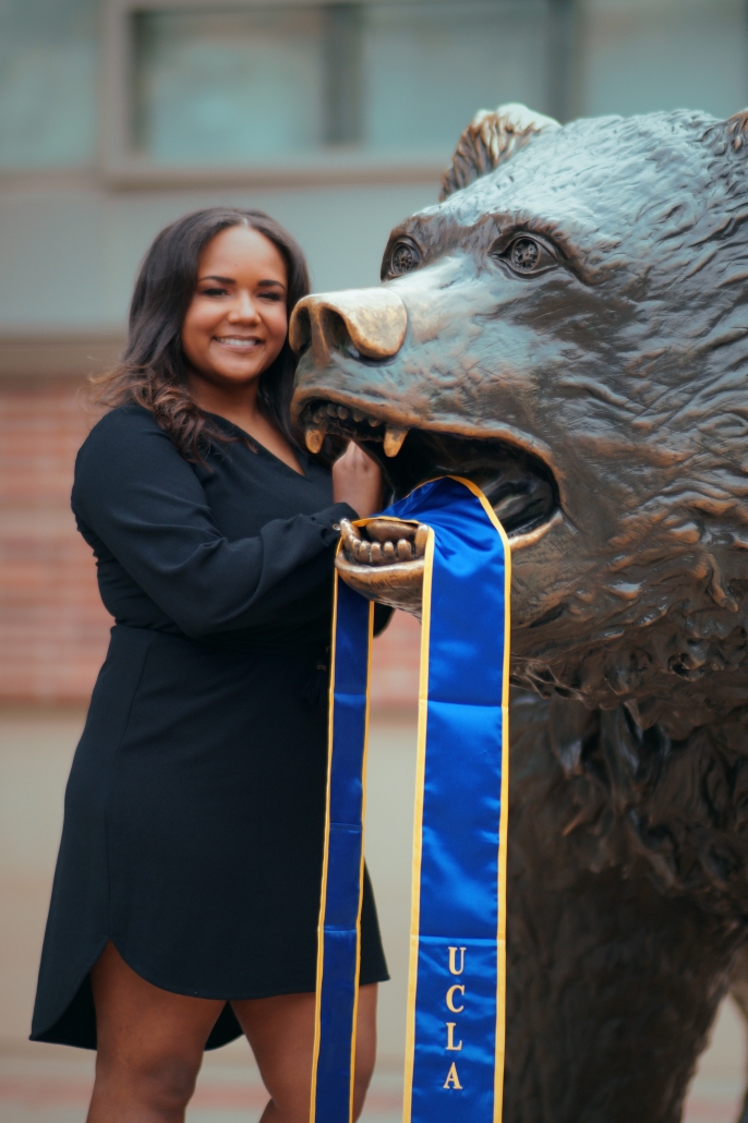 Cynamon Mantley smiles beside UCLA’s Bruin bear statue, with her blue-and-gold graduation stole draped through the statue’s mouth.
