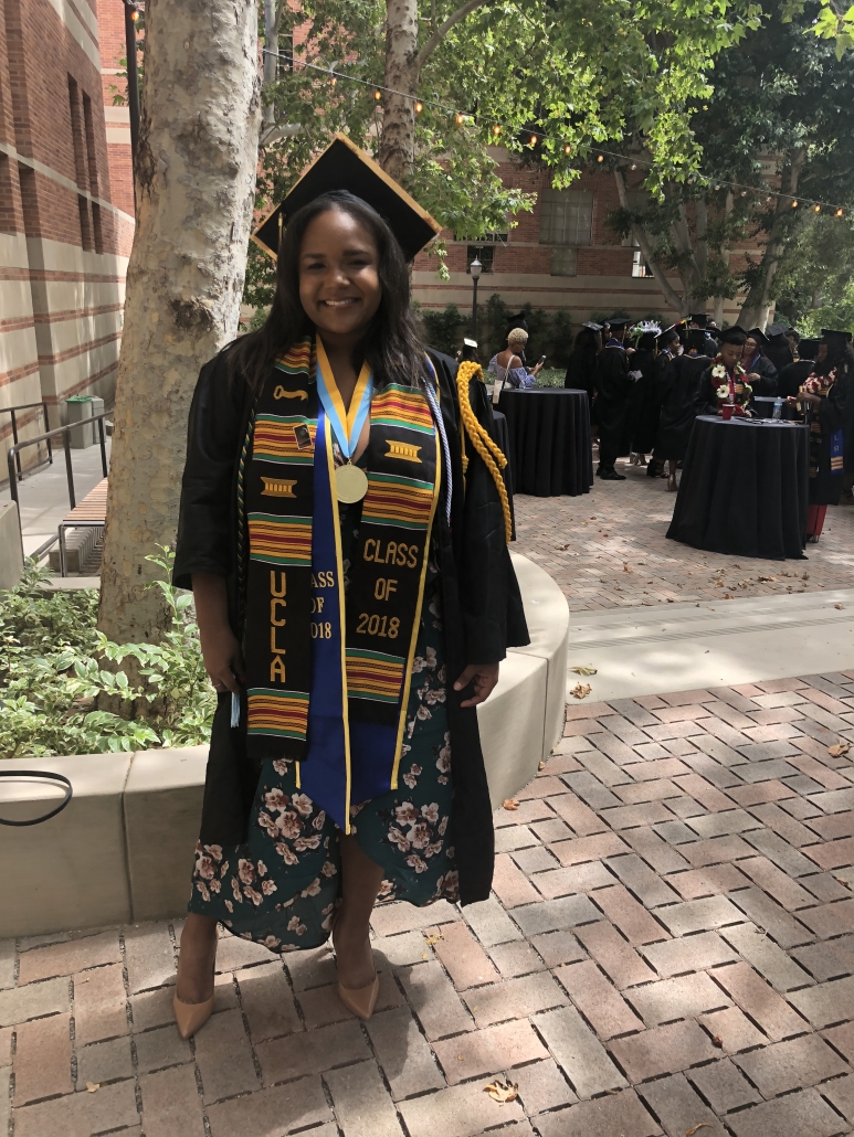 Cynamon Mantley ’18 smiles in graduation regalia near a tree, wearing a decorated stole, medal, and blue-and-gold UCLA sash.